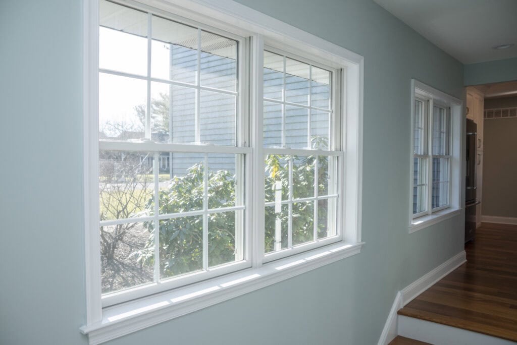A hallway with light blue walls and wooden floors features large white-framed windows, allowing sunlight to enter and providing a view of green bushes and part of a house outside.