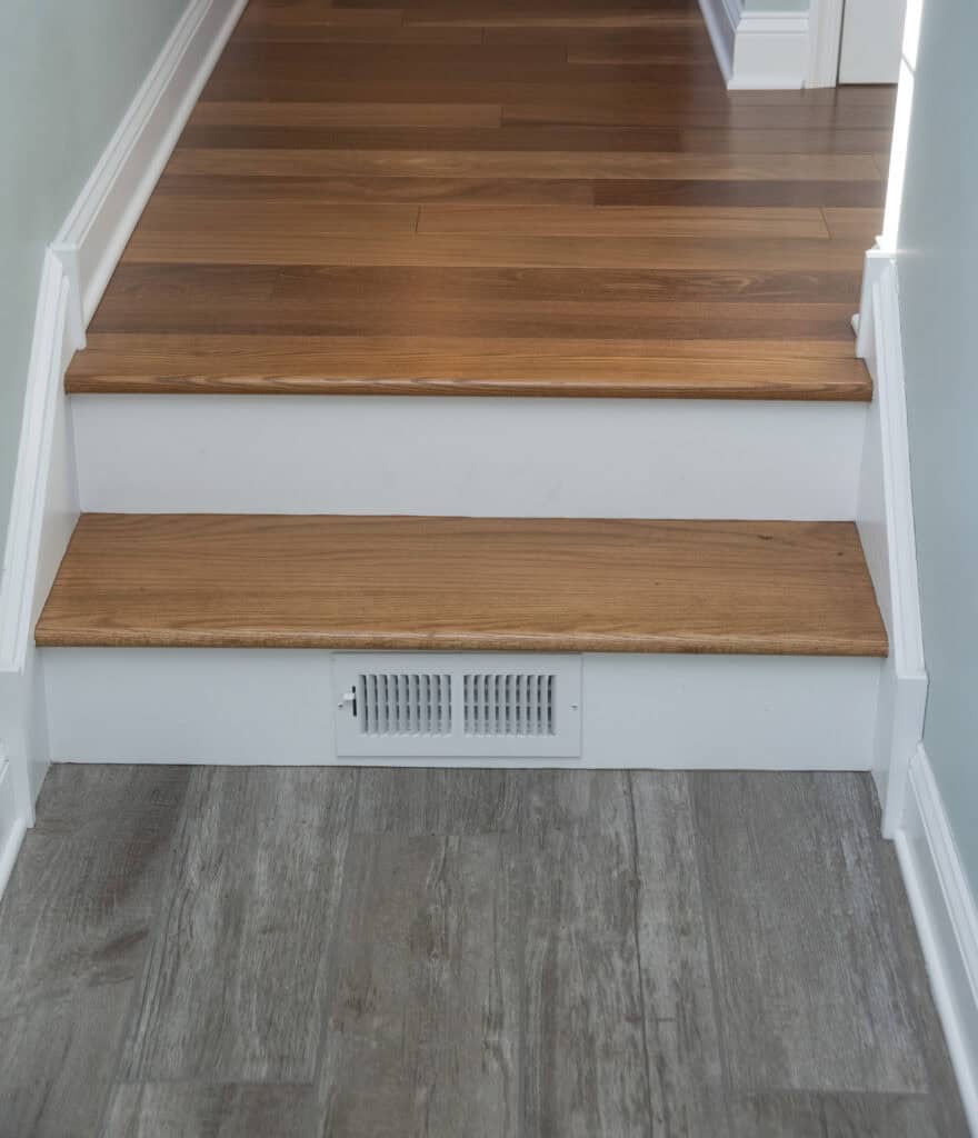 A set of two wooden stairs transitions from tile flooring to hardwood flooring, with a white air vent installed in the riser of the bottom step.
