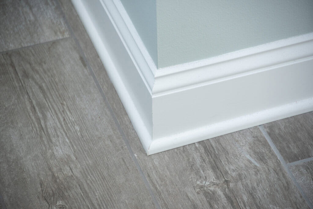 A close-up view of a light-colored baseboard along a corner where two walls meet, with light wood-look tile flooring beneath. The baseboard is clean and has a smooth, glossy finish.