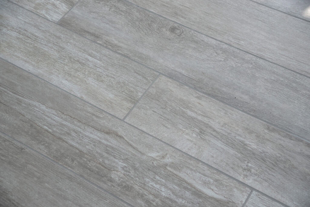Close-up of a floor with rectangular, gray tiles arranged in a staggered pattern. The tiles have a wood grain texture and subtle color variations, resembling weathered wood planks.