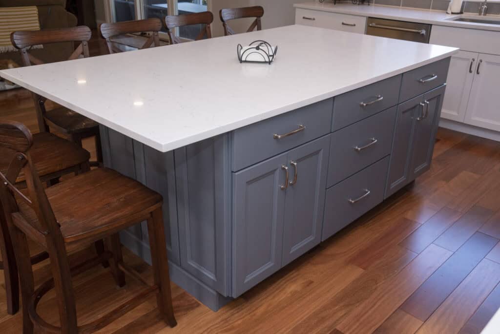 A kitchen island with a white countertop and blue cabinets, surrounded by wooden chairs on a hardwood floor. A small decorative item sits on the counter.