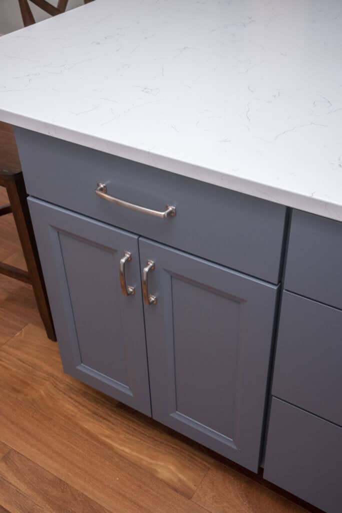 Blue kitchen cabinets with silver handles and a white marble countertop, set on a wooden floor. A dark wooden chair is partially visible next to the cabinets.