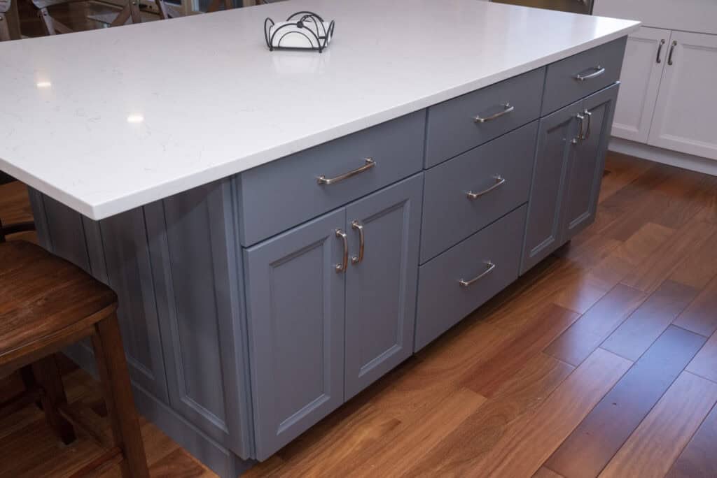 A kitchen island with light blue cabinets, silver handles, and a white marble countertop, set on a wooden floor. A wooden stool and metal napkin holder are also visible.