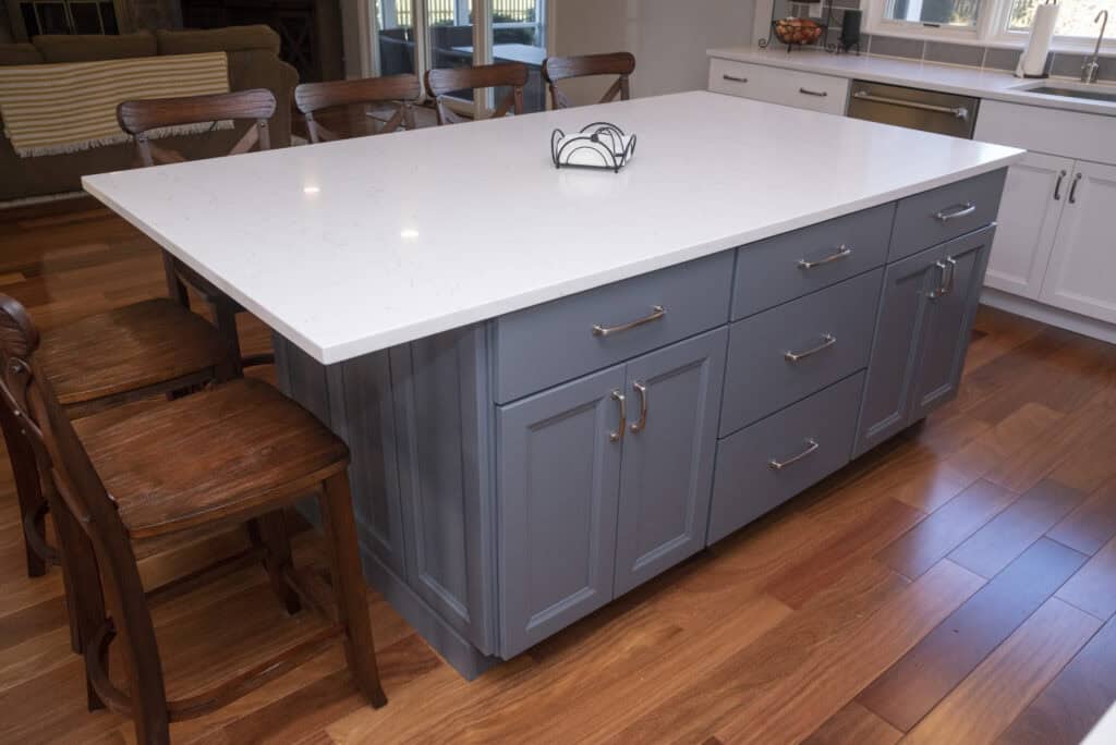 A modern kitchen island with a white marble countertop, gray-blue cabinets, silver handles, and wooden chairs on a hardwood floor. A small metal napkin holder sits on the counter.