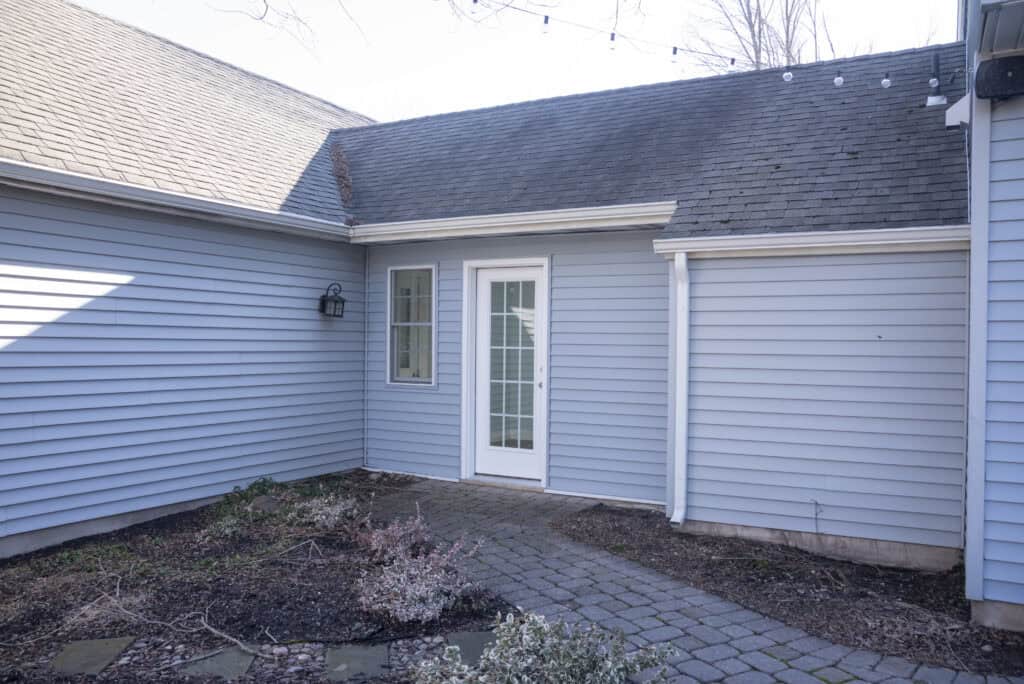 A small patio with gray siding walls, a glass-paneled door in the center, and a side window. There are some garden beds with sparse plants and a stone walkway in front of the entrance.