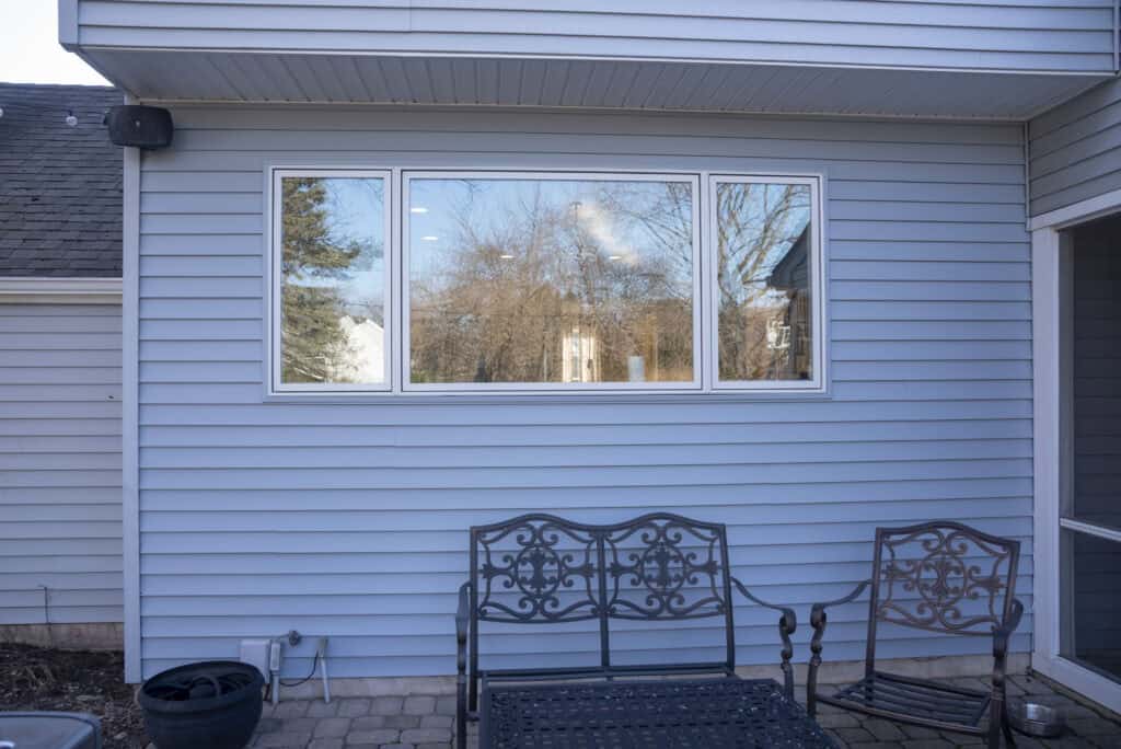 Three decorative metal chairs sit on a stone patio in front of a blue horizontal siding house wall with three large windows reflecting trees and the sky.