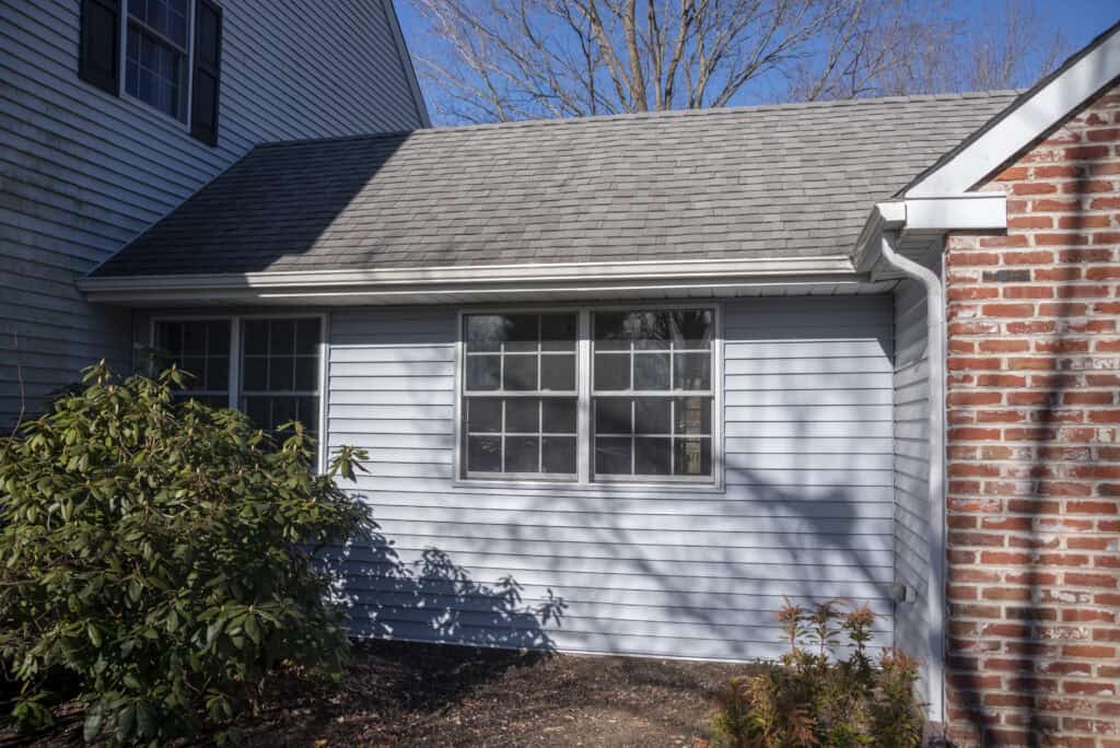 A side view of a house showing light blue siding, a window with divided panes, a brick wall corner, and some shrubs and plants in the foreground under a clear blue sky.