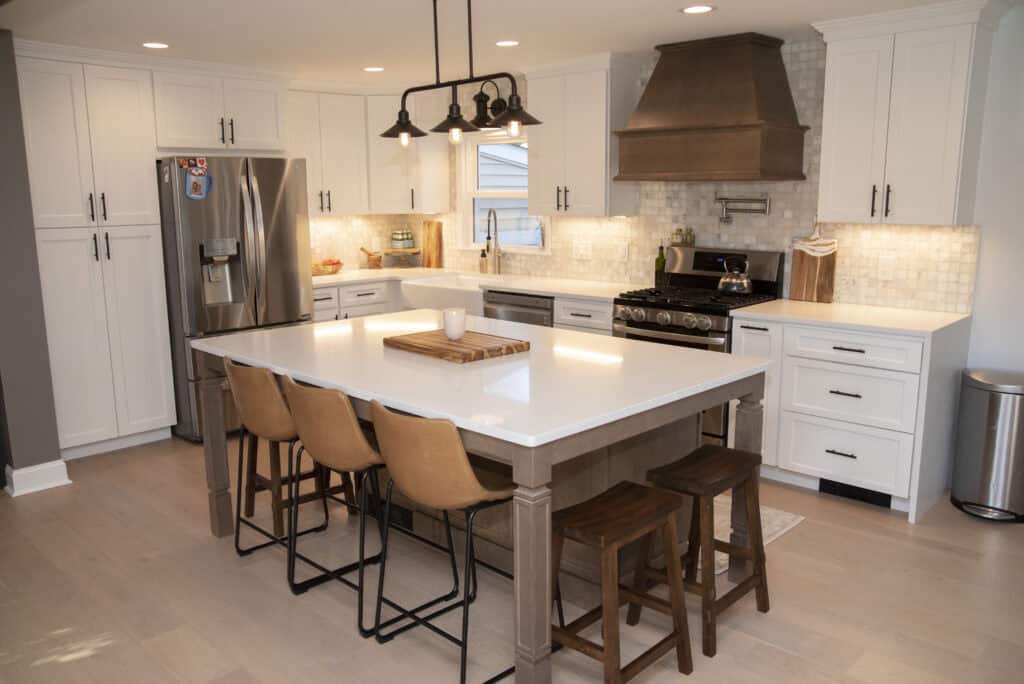 Modern kitchen with white cabinets, stainless steel appliances, a large center island with four tan barstools, pendant lighting, and a brown range hood over a gas stove. Light wood flooring completes the space.