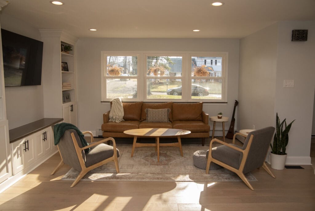 A cozy living room with a brown leather sofa, two gray armchairs, a round wooden coffee table, and large windows letting in natural light. Decorative plants and bookshelves accent the space.