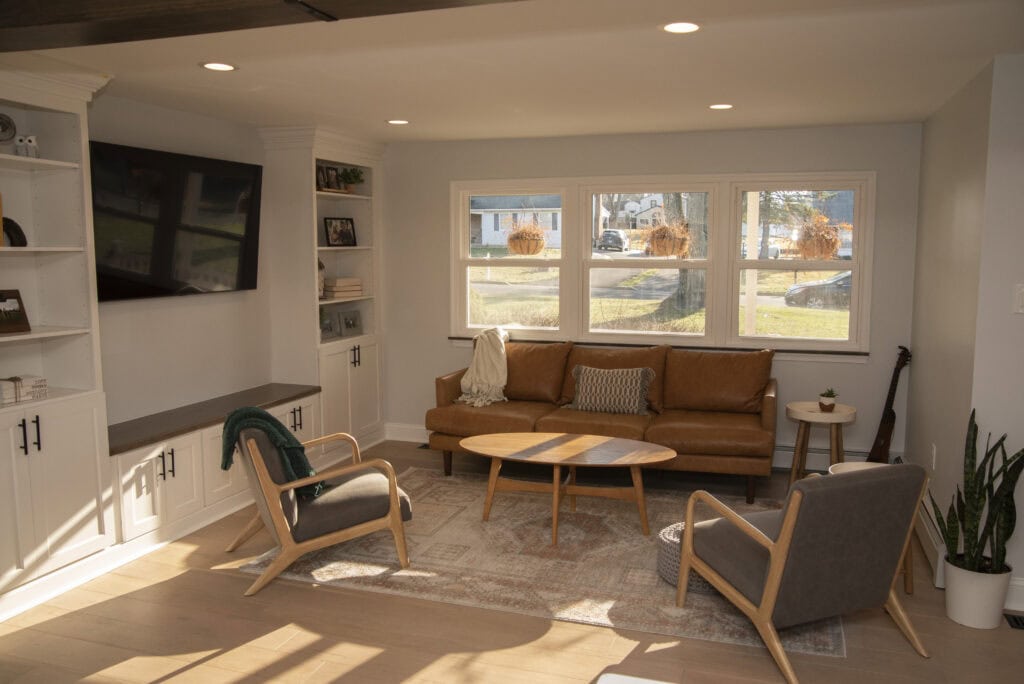 A modern living room with a tan leather sofa, two gray armchairs, a wooden coffee table, built-in white shelves, a wall-mounted TV, large windows, and sunlight streaming in.