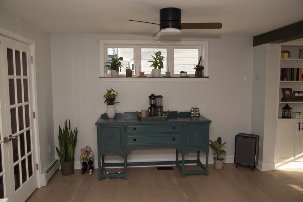A teal sideboard with a coffee maker and flowers stands against a white wall beneath a row of small windows with potted plants. The room has light wood floors, a ceiling fan, and a French door.