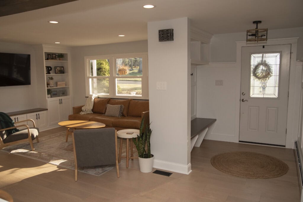 A cozy living room with a brown leather sofa, two chairs, a round wooden coffee table, and a potted plant. Near the entryway is a white door with a wreath and a round rug. Sunlight streams in through large windows.