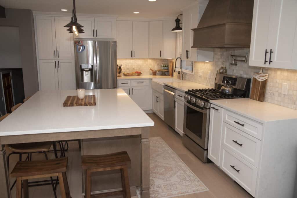 Modern kitchen with white cabinets, stainless steel appliances, a large island with wooden stools, pendant lighting, and a rug on the light wood floor. A refrigerator, stove, and sink are visible.