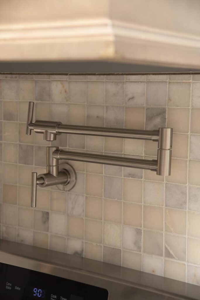 A stainless steel pot filler faucet is mounted on a tiled kitchen backsplash above a stove. The faucet has an articulated arm for extending and folding.