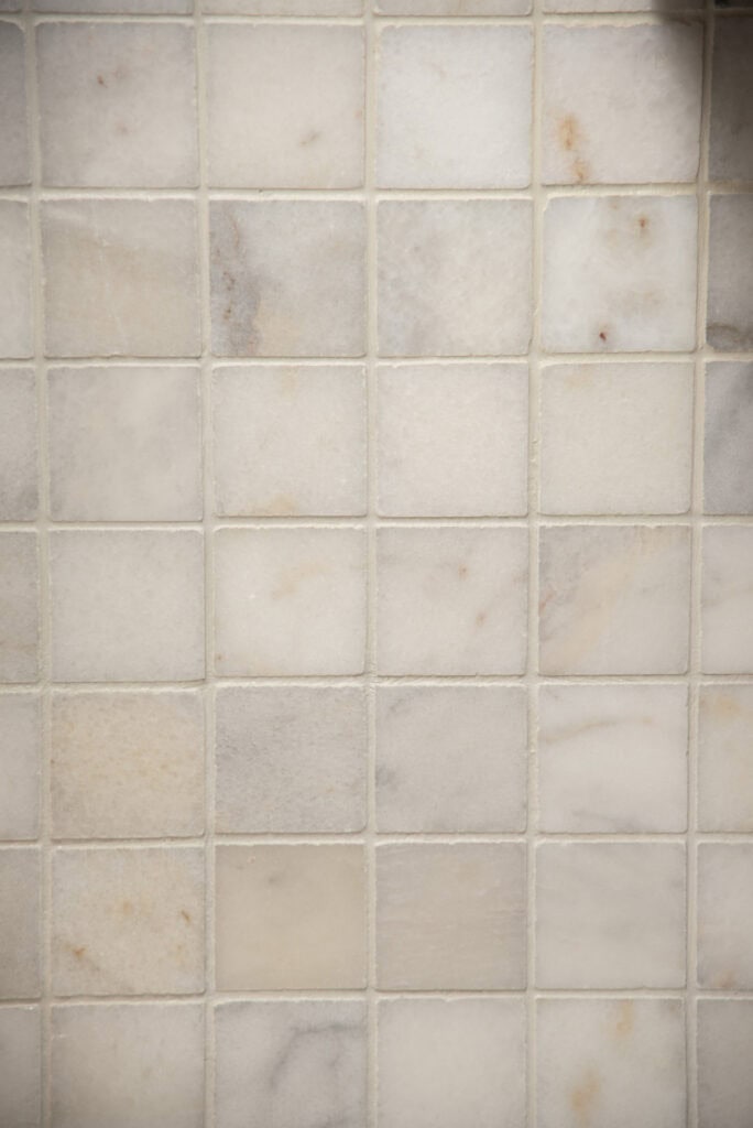 Close-up of a tiled surface with small, square tiles in light beige and off-white shades, featuring subtle natural stone patterns and variations. The grout lines are thin and light-colored.