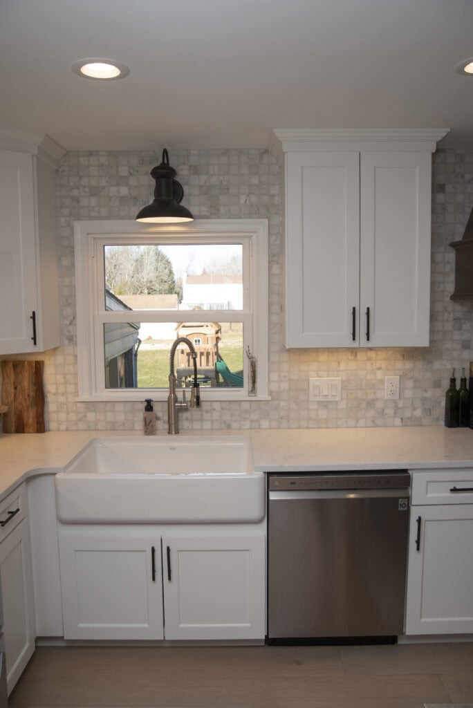 A modern kitchen with white cabinets, a farmhouse sink, stainless steel dishwasher, and a window above the sink overlooking a backyard with a playset. The backsplash is made of small, light-colored tiles.