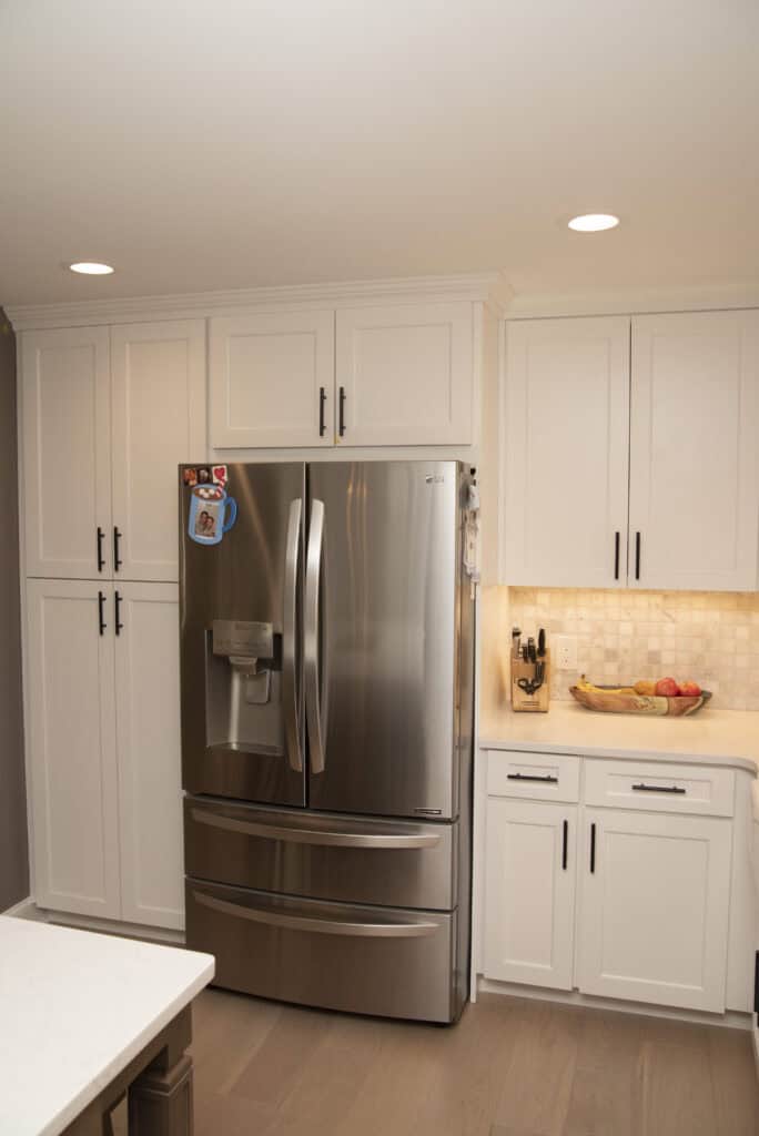 A modern kitchen with white cabinets, a stainless steel French door refrigerator, some magnets on the fridge, a countertop with a fruit bowl, and a knife block under warm recessed lighting.