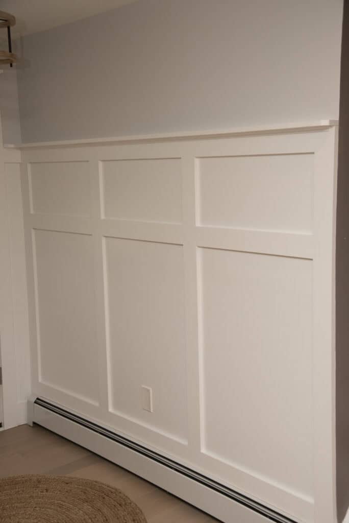 A section of an interior wall features white board and batten paneling, a light gray upper wall, a round tan rug on light wood flooring, and a white baseboard heater with a visible electrical outlet.