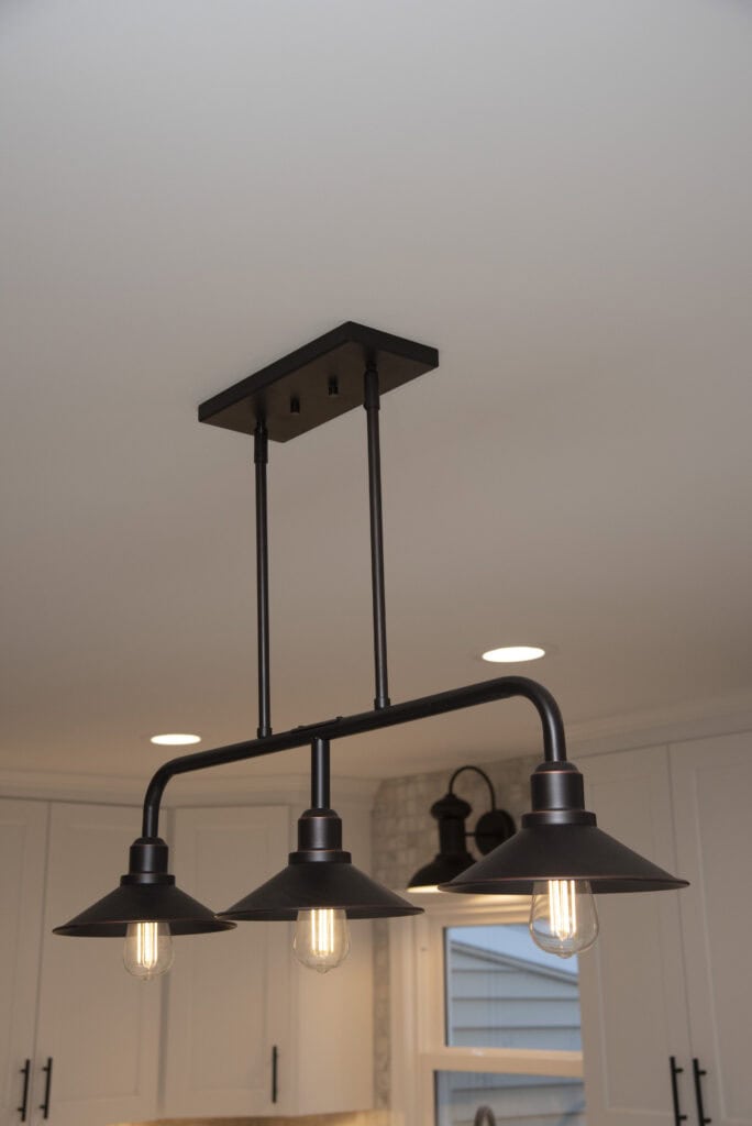 A black metal ceiling light fixture with three downward-facing pendant lamps and exposed bulbs, mounted on a white ceiling in a modern kitchen.