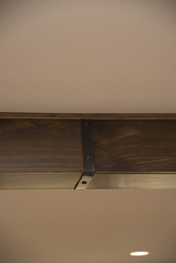 A close-up of a wooden ceiling beam with a dark metal bracket attached, set against a beige ceiling. A small round recessed light is partially visible at the bottom right corner.