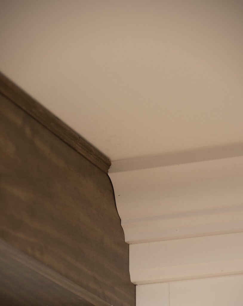 Close-up of a ceiling corner showing a joint between white crown molding and dark wood trim with a visible gap, highlighting an imperfect fit.