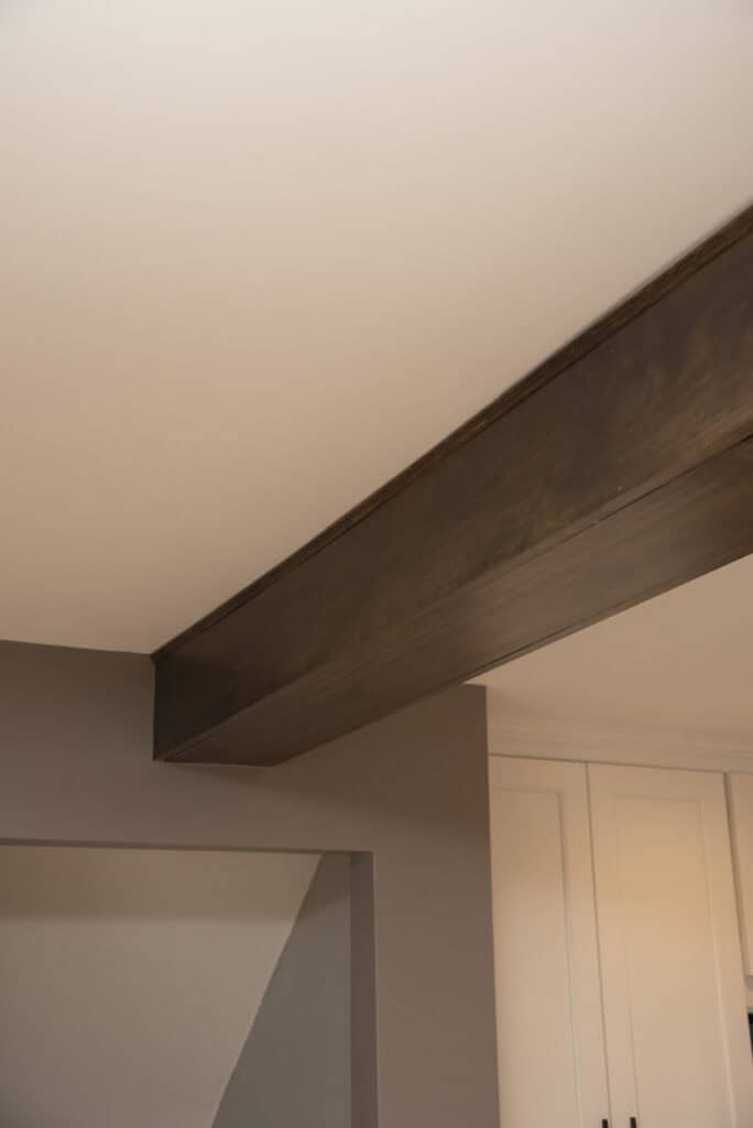 A dark wooden ceiling beam runs across a light-colored ceiling in a room with beige walls and white cabinets in the background.
