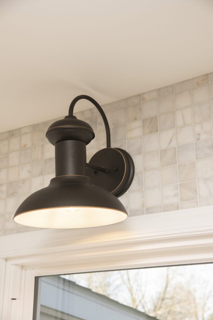 A black metal wall sconce with a wide shade is mounted on a tiled wall above a window, casting light downward. The tiles are light-colored and have a subtle grid pattern.