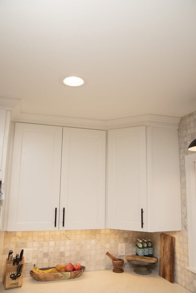 White kitchen cabinets with black handles are mounted on the wall above a countertop. On the counter, there is a bowl of fruit, a knife block, some bottles, a mortar and pestle, and a wooden cutting board.