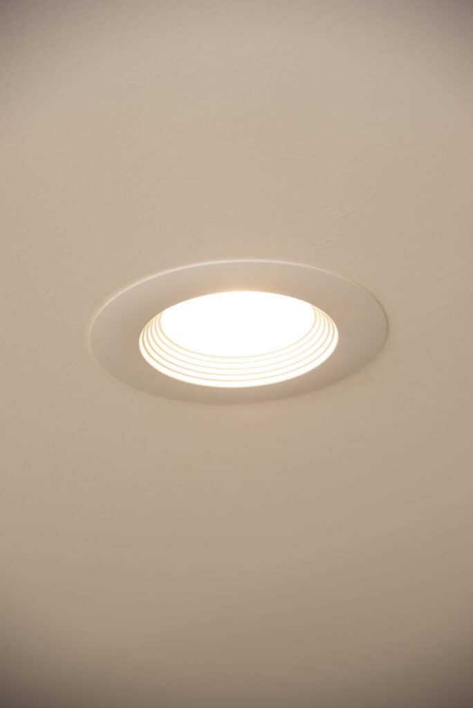 A close-up view of a recessed ceiling light turned on, set in a white ceiling with concentric ridges around the bulb.