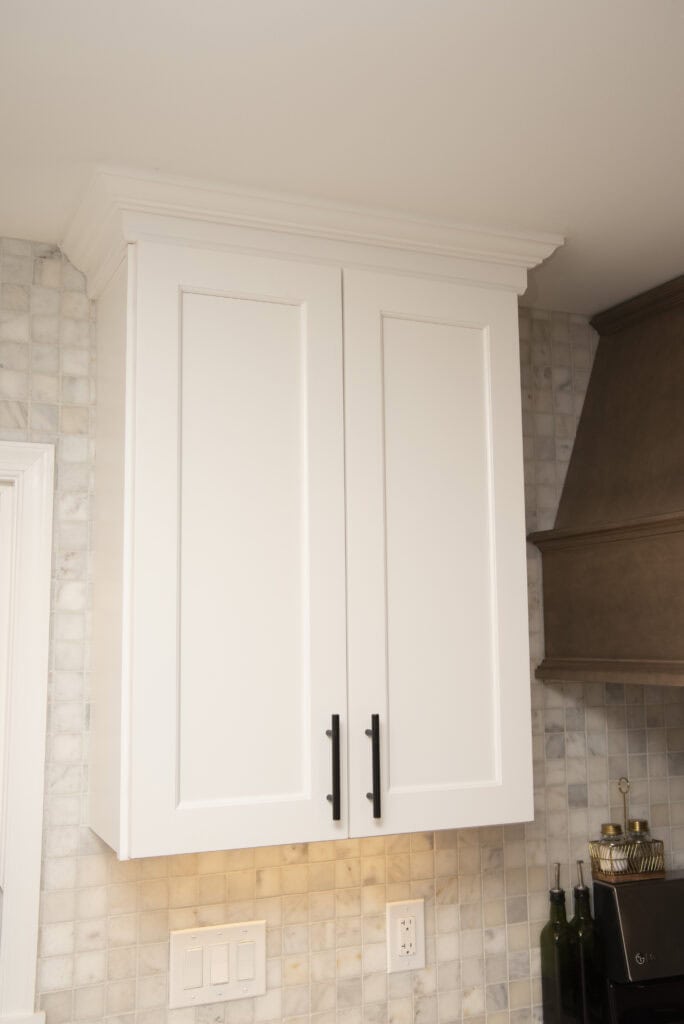 White kitchen cabinet with two doors and black handles mounted on a wall with light-colored tile backsplash; a range hood and bottles are visible on the right side.