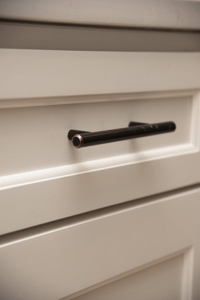 Close-up of a white cabinet drawer with a modern black metal handle attached horizontally at the center. The cabinet has a raised panel design and a smooth, clean finish.