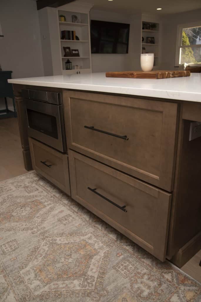 A kitchen island with brown cabinets, black handles, a built-in microwave, and a white countertop. A wooden tray with a candle sits on top. Patterned rug partially visible on the floor. Shelves and TV in the background.