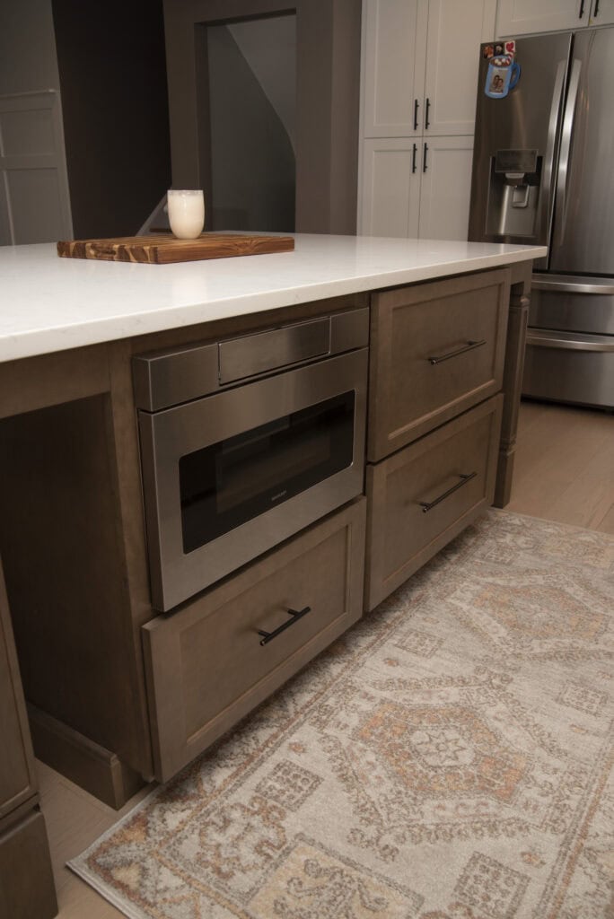 A modern kitchen features a built-in microwave and large drawers in a brown island, a white countertop with a candle on a wooden tray, patterned rug, and stainless steel refrigerator in the background.