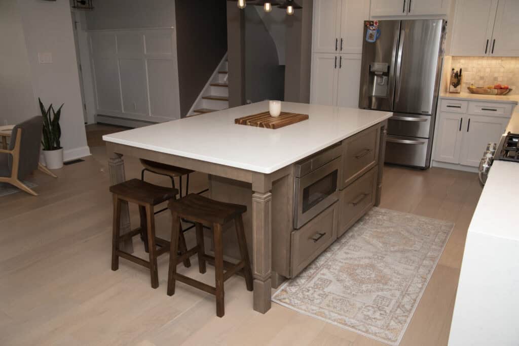 A modern kitchen with a large island featuring a white countertop, two wooden stools, a wooden cutting board, and a candle. Stainless steel appliances and a light-colored rug are also visible.