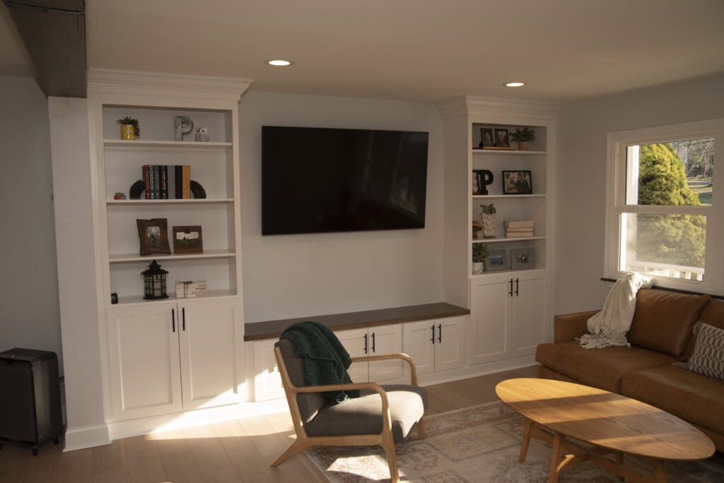 A cozy living room with a wall-mounted TV between two white built-in bookshelves, a tan leather sofa, a wooden chair with a green blanket, a round wooden coffee table, and sunlight streaming through a window.