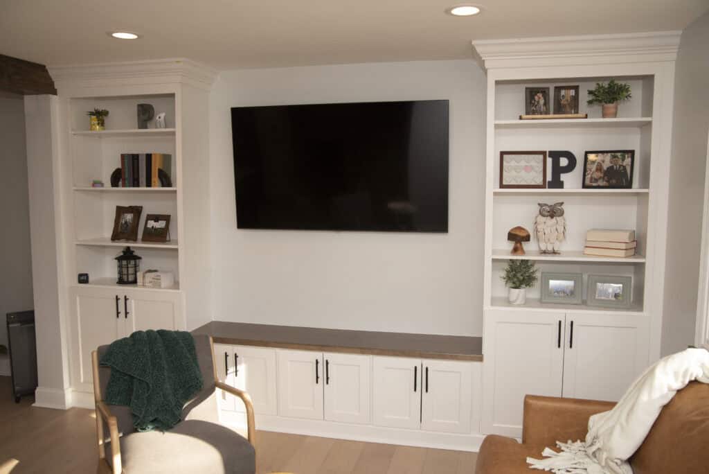 A living room with built-in white shelves and cabinets on either side of a wall-mounted flat-screen TV. The shelves display books, framed photos, plants, and decorative objects. A brown chair is partially visible in the foreground.