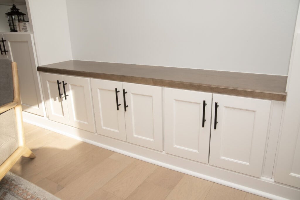 Built-in white cabinets with paneled doors and black handles, topped with a wooden countertop. The cabinets are set against a white wall on a light wood floor, with part of a chair and shelf visible at the edges.