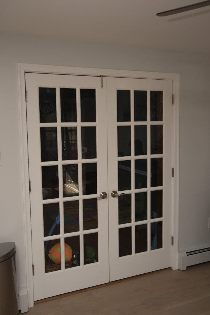 White French doors with glass panes reveal a glimpse of a playroom inside, including a colorful ball and toys on the floor. The doors have silver handles and are set in a light-colored wall.