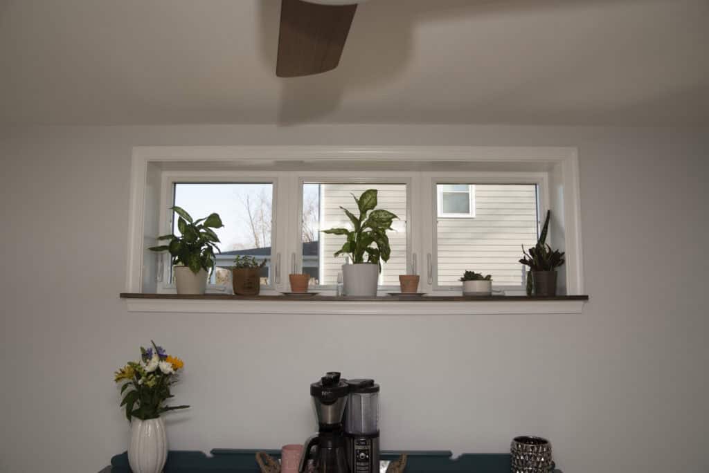 A wide window sill holds five potted plants; outside, a house and a leafless tree are visible. Below the sill sits a coffee maker and a vase with yellow, white, and purple flowers on a teal table.