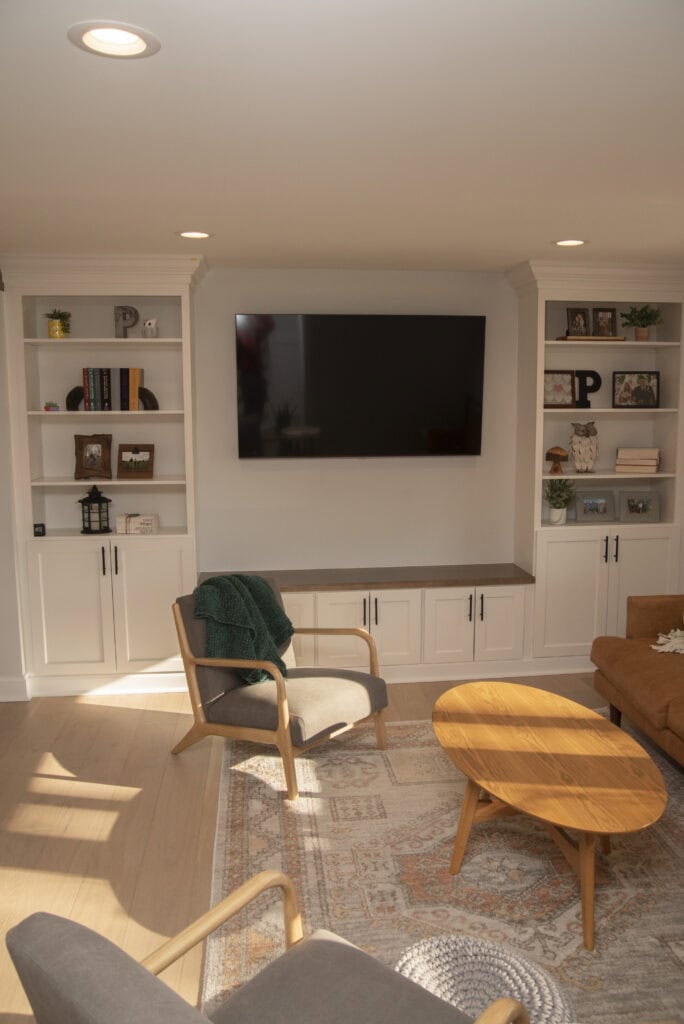 A cozy living room with built-in white shelves, a wall-mounted TV, two wooden armchairs, a round wooden coffee table, and a patterned rug. Sunlight streams in through a window, brightening the space.