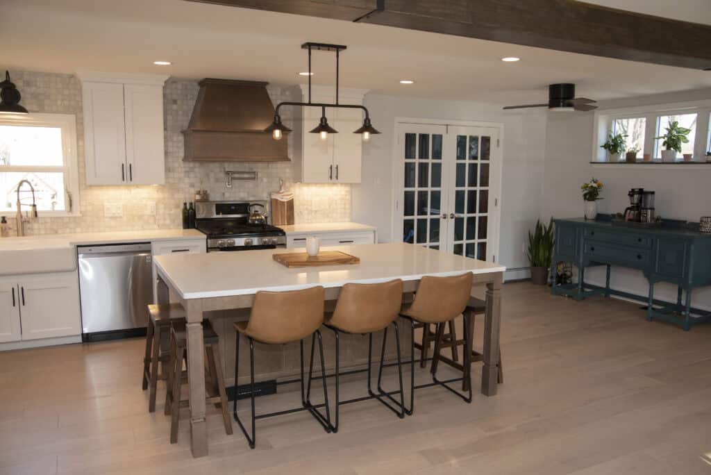 Modern kitchen with a large island featuring four tan barstools, white cabinets, stainless steel appliances, a range hood, pendant lights, and a blue sideboard against the wall.