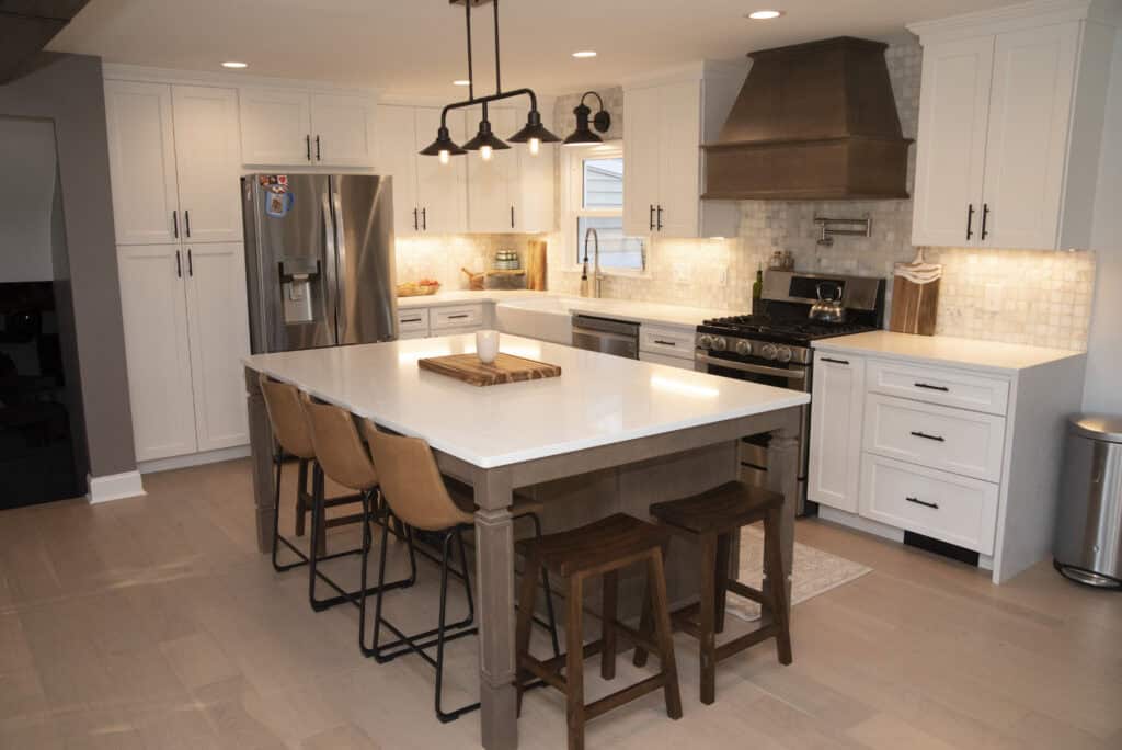Modern kitchen with white cabinets, stainless steel appliances, a large island with wooden stools, pendant lights above, and light wood flooring. The backsplash is tiled and countertops are white.
