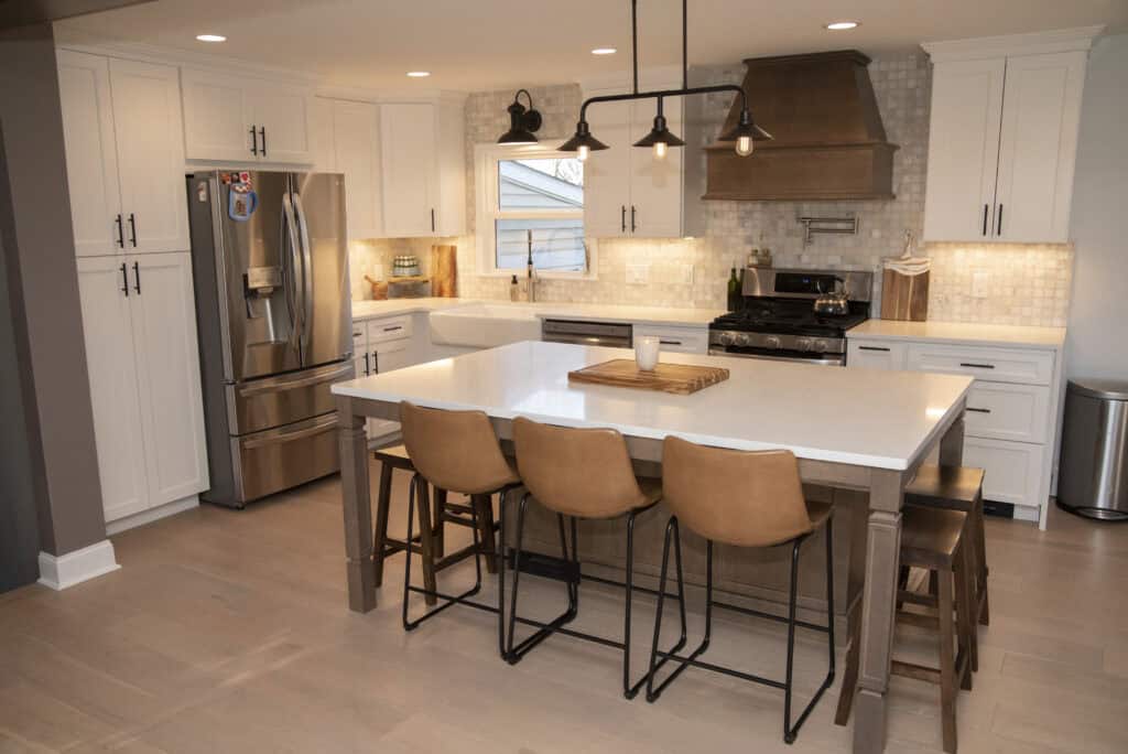 Modern kitchen with white cabinets, stainless steel appliances, a large island with a white countertop, four brown bar stools, pendant lights, and a tile backsplash behind the stove and range hood.
