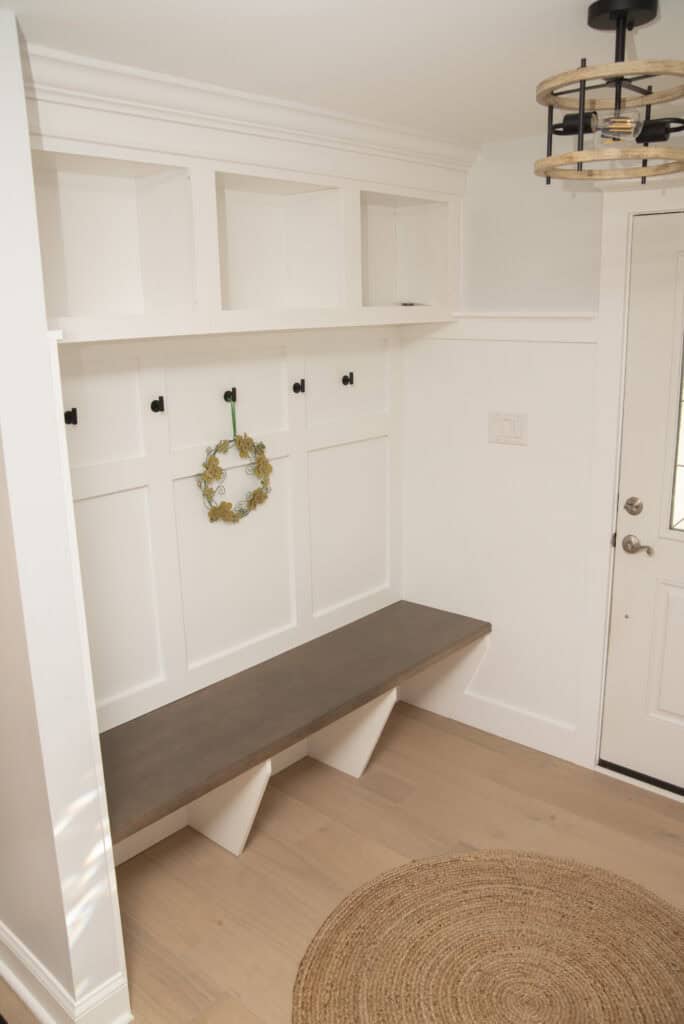 A mudroom with white built-in cubbies, black hooks, a wooden bench, a small green wreath hanging on one hook, a round woven rug, and a white door with glass panels.