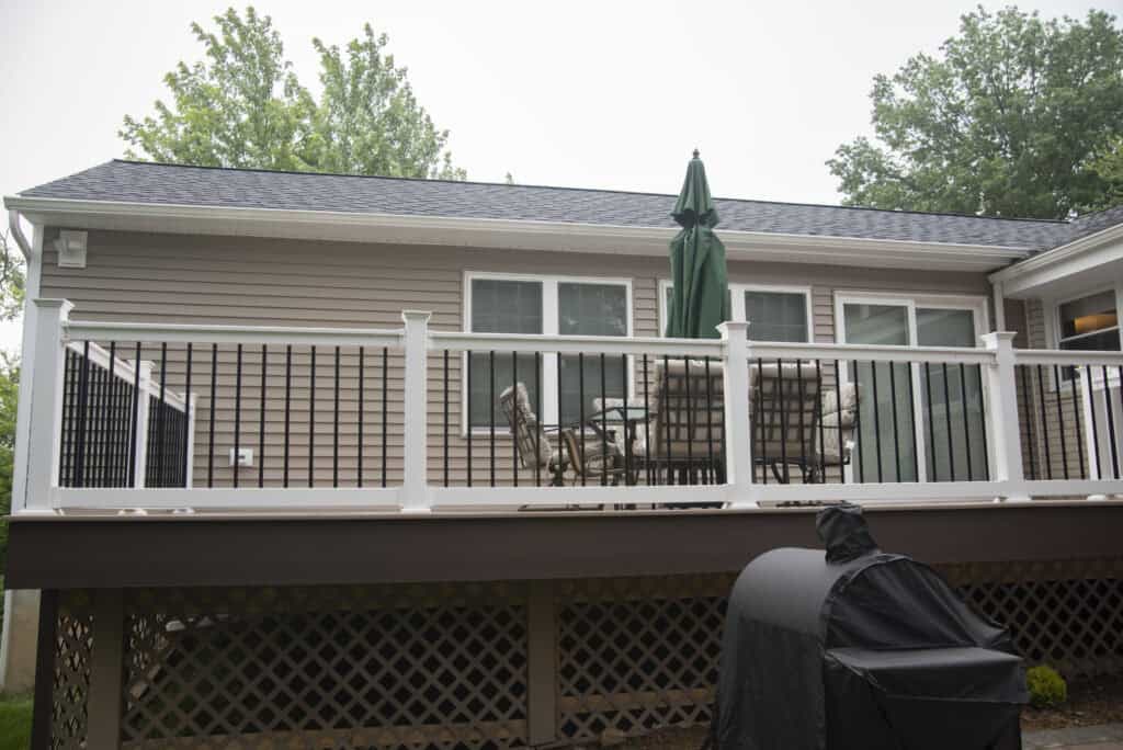 A backyard wooden deck attached to a beige house, with black-and-white railing, a patio table with chairs, and a closed green umbrella. A covered grill is visible in the foreground. Trees are in the background.