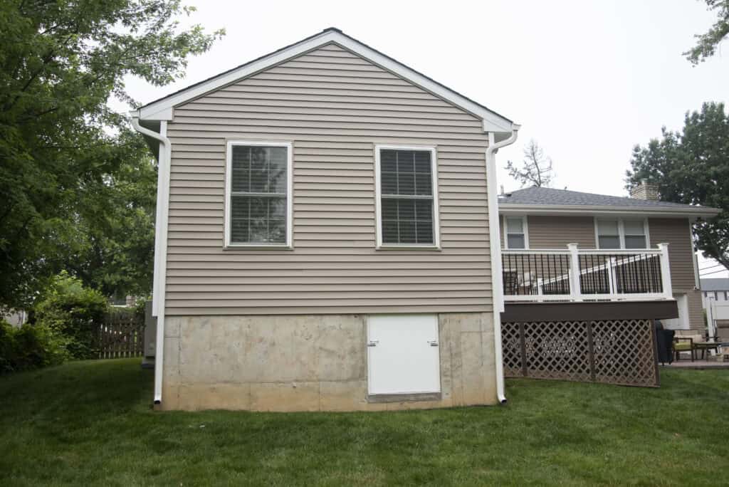 A beige house with white trim, two windows, and a small white door at the foundation level, sits on a grassy lawn. There is a wooden deck with white railing attached to the right side of the house.