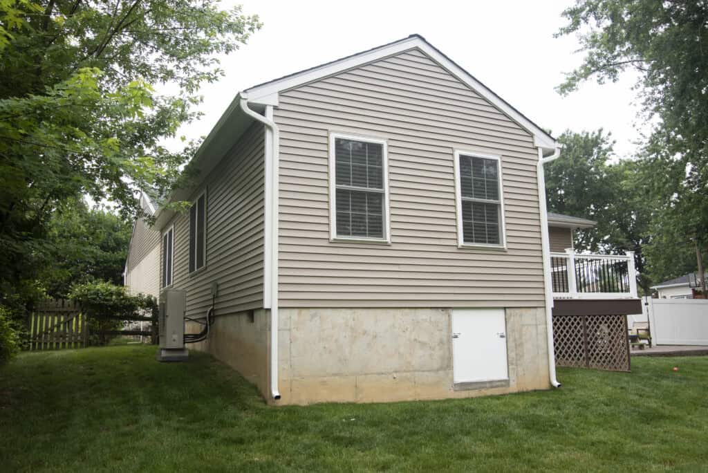 A beige house with vinyl siding, two windows, white trim, and a small white door on a concrete foundation, surrounded by green grass and trees. A wooden deck is attached to one side of the house.