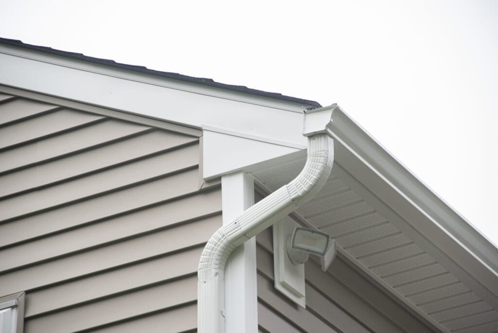 Close-up of the corner of a house showing beige vinyl siding, a white gutter system with a downspout, and a security light mounted under the eaves against a cloudy sky.