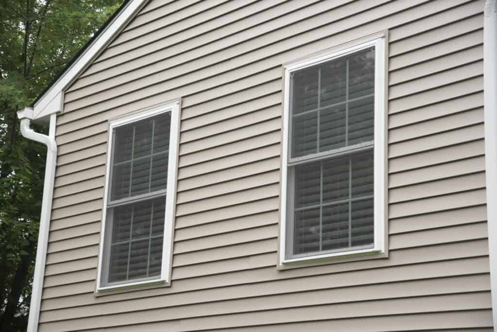 A beige house exterior with vinyl siding and two white-framed double windows, each with closed blinds. A white gutter runs along the roofline, and green tree branches are visible in the upper left corner.