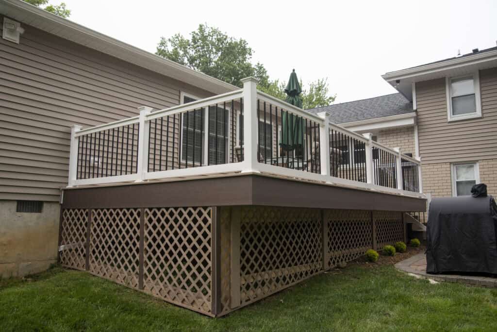 A raised backyard deck with white railings and brown lattice skirting, attached to a beige house. Patio furniture with a green umbrella sits on the deck, and a grill is nearby on a stone patio. Grass surrounds the area.